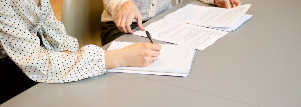 woman signing on white printer paper beside woman about to touch the documents