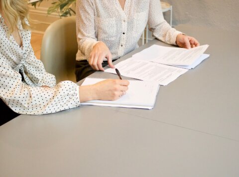 woman signing on white printer paper beside woman about to touch the documents