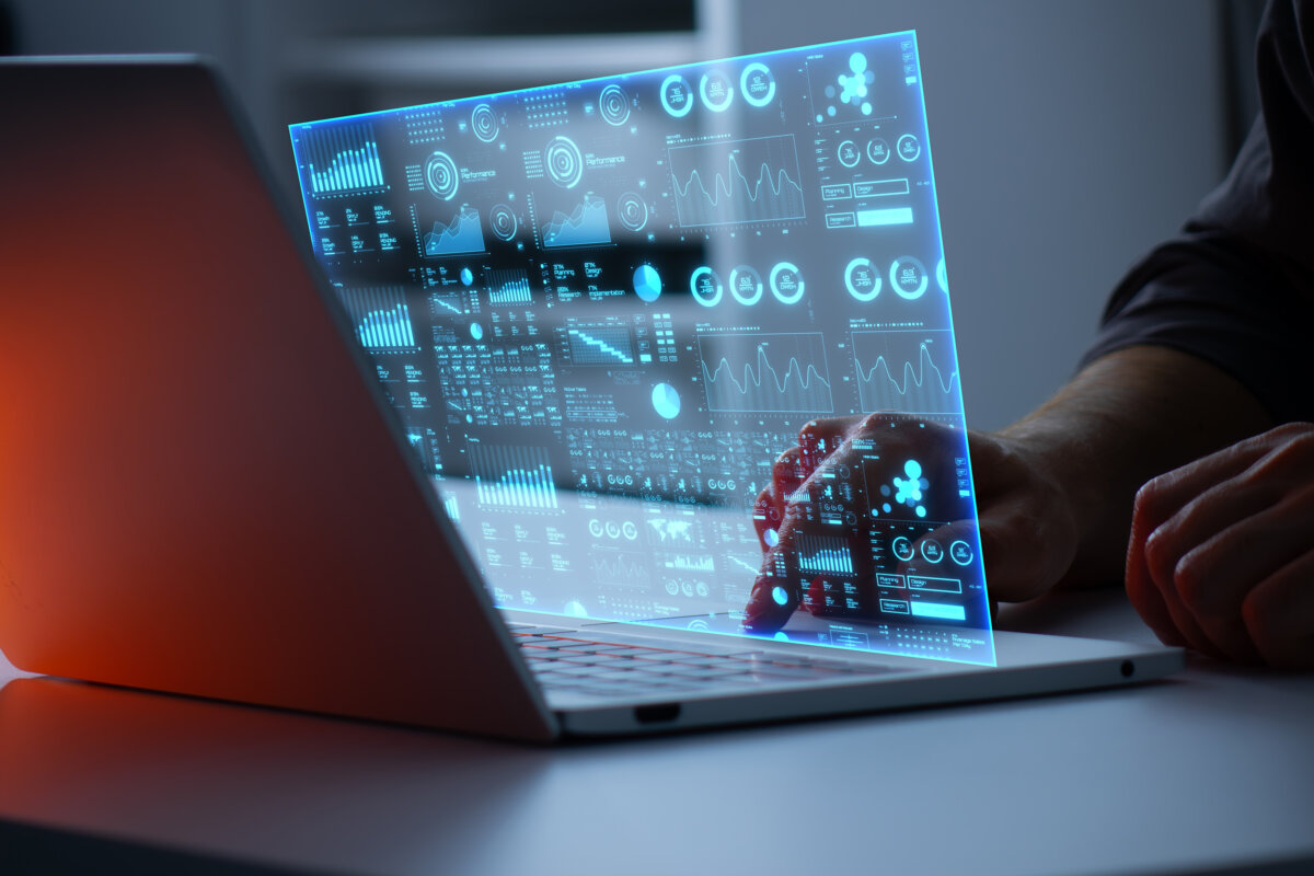 Holographic blue data dashboard hovering above an open laptop, held by a person in viewable silhouette.